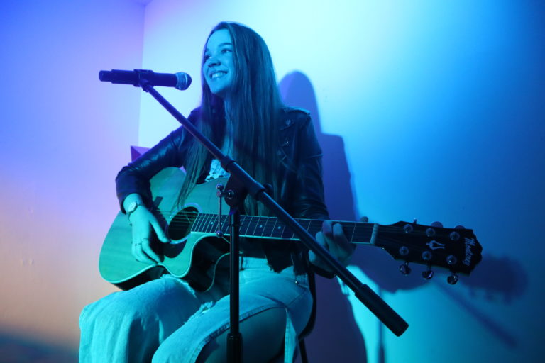 young girl holding guitar and singing into microphone smiling under blue and purple light