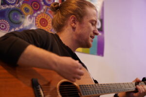 Talented Coach Liam strums acoustic guitar in inclusive Ballarat music studio.