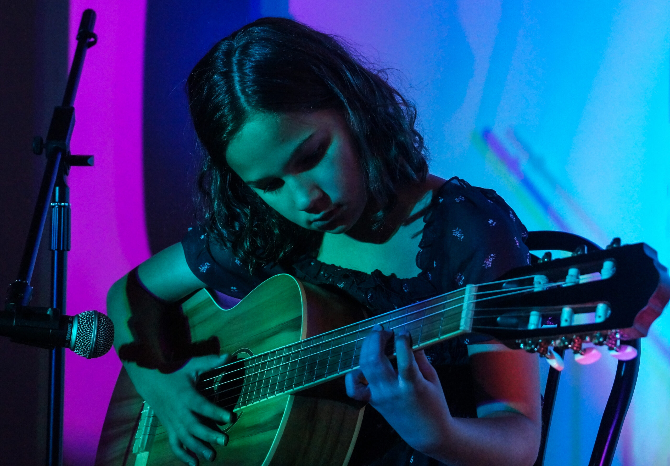 SoundCheck Studios student performs on stage under purple and blue lights with her guitar at First Steps Performance 2024