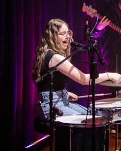Talented drum and vocal student performs on stage with Fifth Layer Music at Summer Sounds with SoundCheck, 2024